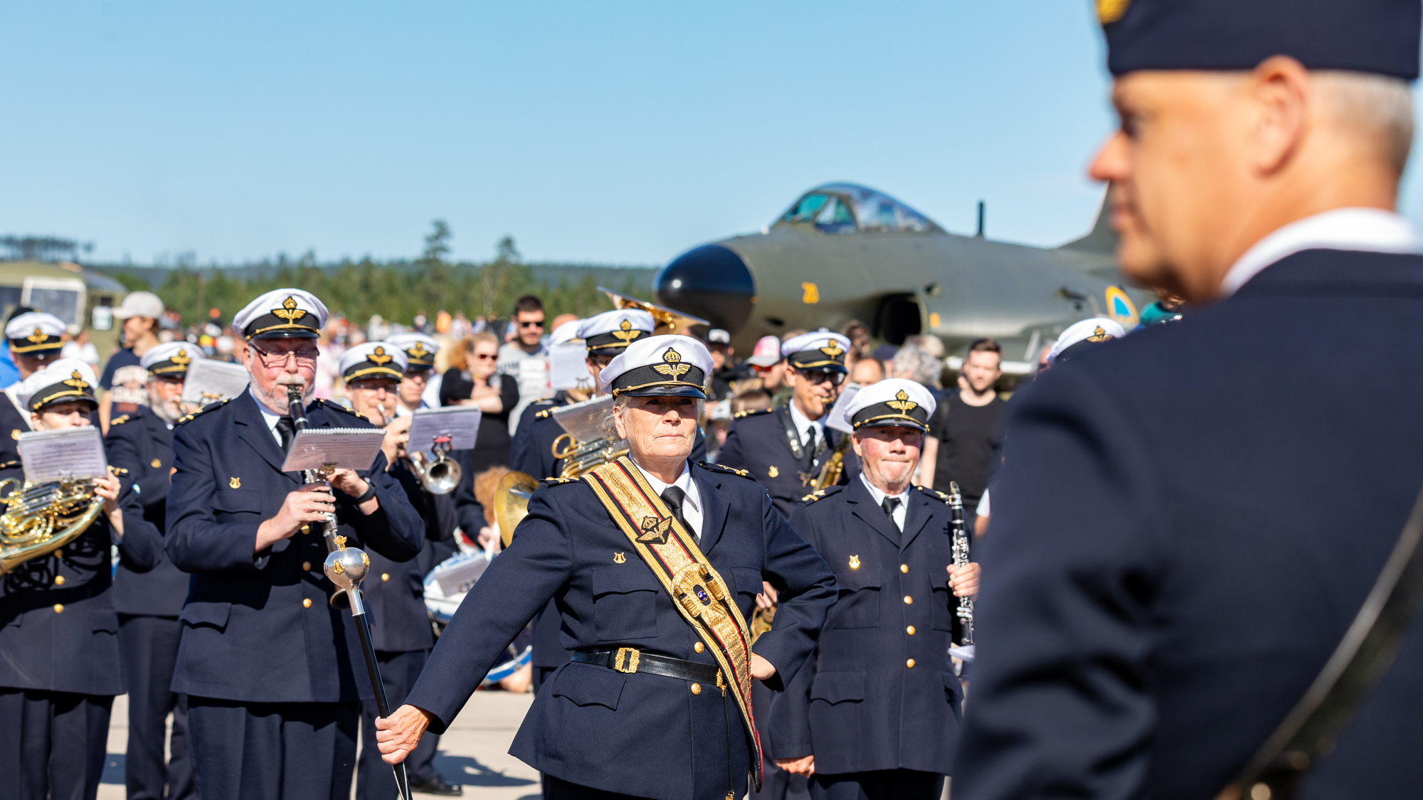 En orkester i uniform spelar p&aring; ett flygf&auml;lt, ett stridsflygplan i bakgrunden och en man i uniform i f&ouml;rgrunden.